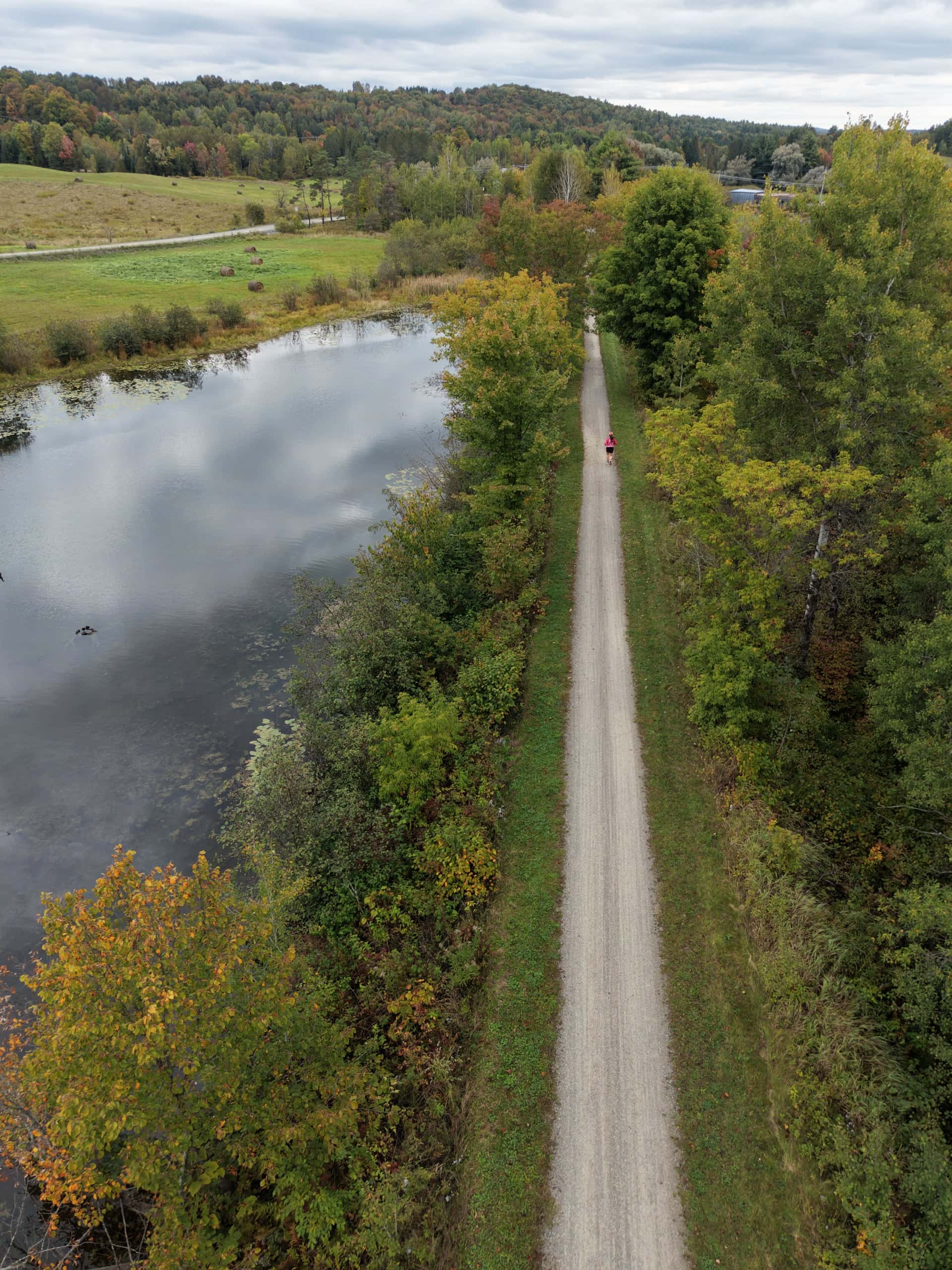 Vue aérienne du Sentier Nature Tomifobia en automne : un chemin de poussière de pierre longe un étang, une coureuse en rose au loin.